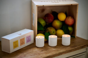 Three lit candles on a wooden surface with a box and a crate of fruits in the background.