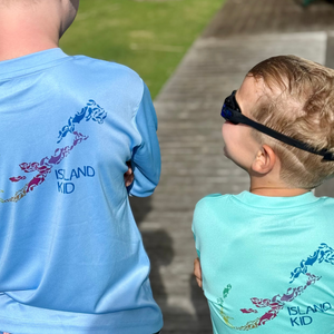 Two children wearing blue 'Island Kid' shirts on a wooden path with grass and trees in the background.