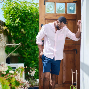 Man in a pink shirt and blue shorts standing in a garden with plants and a wooden door.