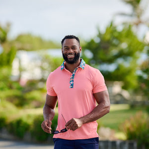 Man wearing a pink polo shirt and navy shorts standing outdoors with greenery in the background