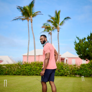 Man in a pink shirt and blue shorts standing on a grassy field with palm trees and buildings in the background.