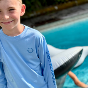 Child wearing a light blue long-sleeve shirt with a logo, standing by a pool with an inflatable shark.