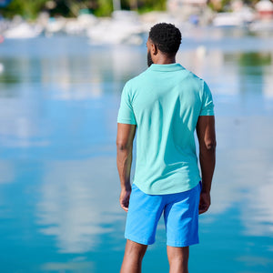 Man in turquoise polo shirt and blue shorts standing on a dock by a body of water.
