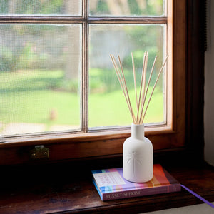 White diffuser with reeds on a windowsill with a view of greenery outside.