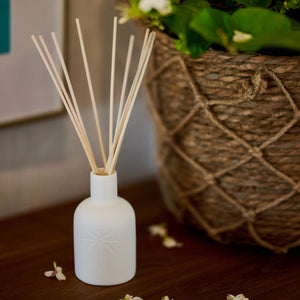 White diffuser bottle with wooden sticks on a wooden surface, with a woven basket and flowers in the background.
