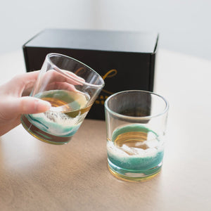 Two glasses with green and white designs on a wooden surface, one being held by a hand.