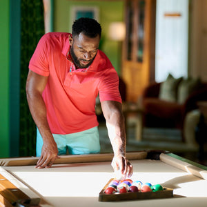 Man in a pink shirt arranging pool balls on a pool table in a living room.