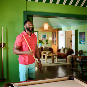 Man in a pink shirt holding a pool cue in a room with green walls and a pool table.