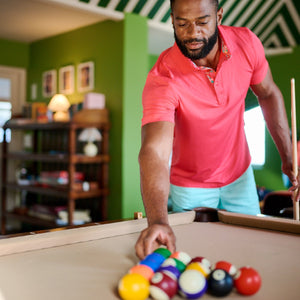 Man in a pink shirt arranging pool balls on a pool table in a room with green walls.