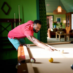 Man in a pink shirt playing pool in a room with green walls and furniture.