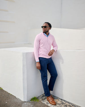 Man in pink sweater and blue pants leaning against a white wall.