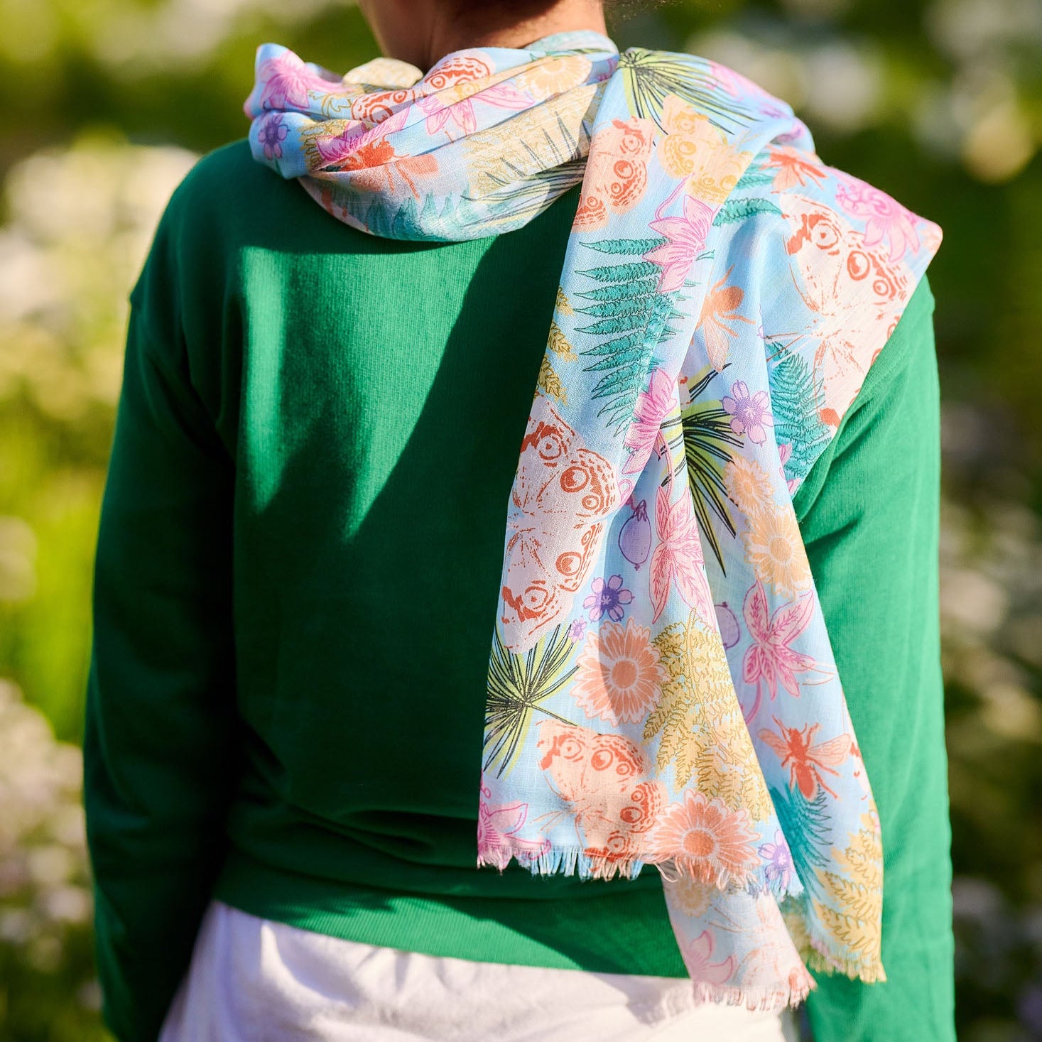 Person wearing a green top and white pants with a colorful floral scarf over their shoulder, standing outdoors.