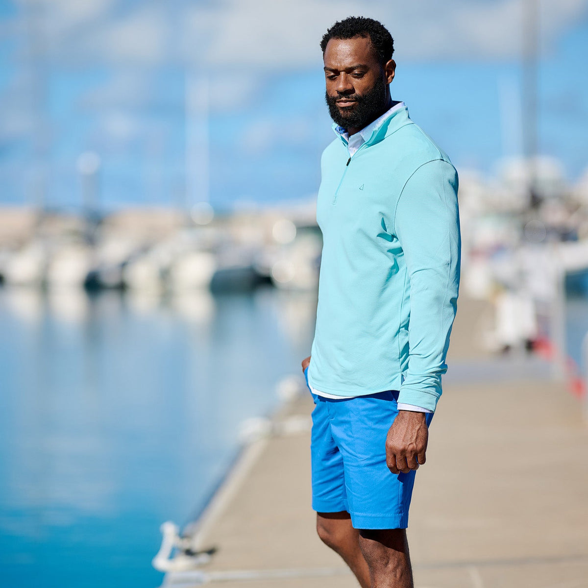 Man wearing a light blue jacket and shorts standing on a dock with water and boats in the background.
