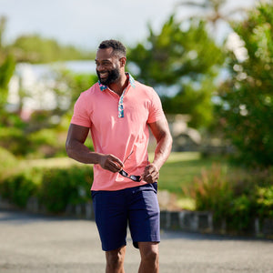 Man in pink polo shirt and navy shorts standing outdoors with greenery in the background
