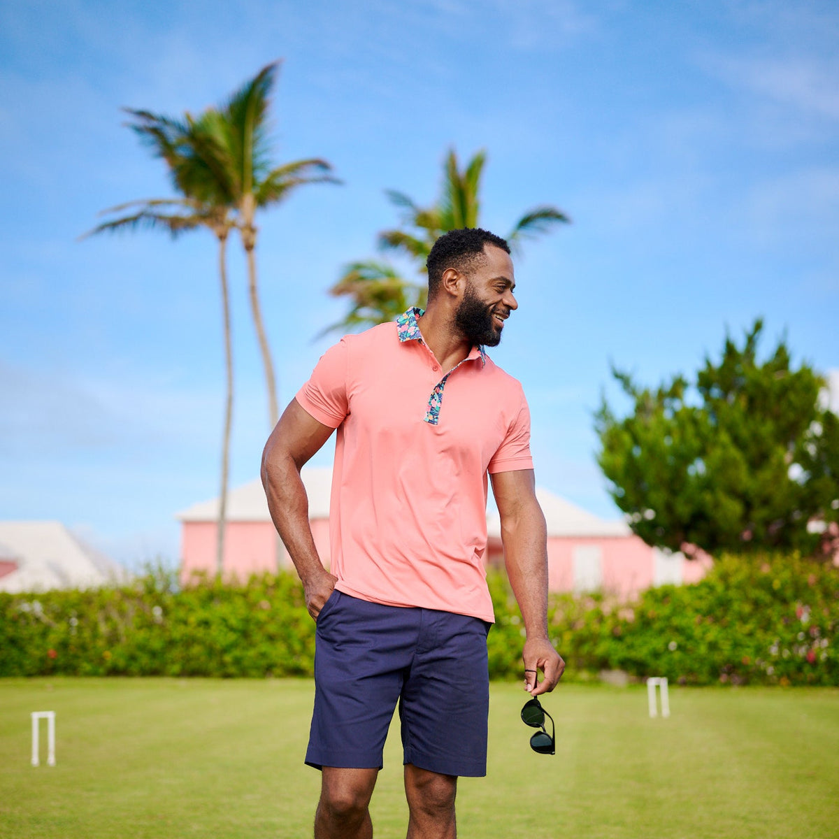 Man in pink shirt and navy shorts standing on a grassy field with palm trees in the background