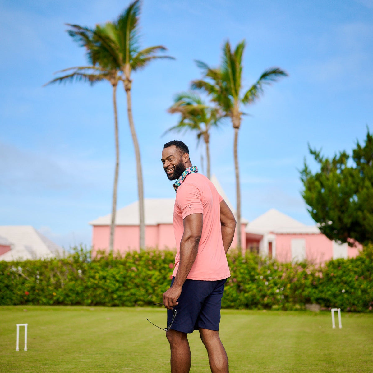 Man in a pink shirt and blue shorts standing on a grassy field with palm trees and buildings in the background.