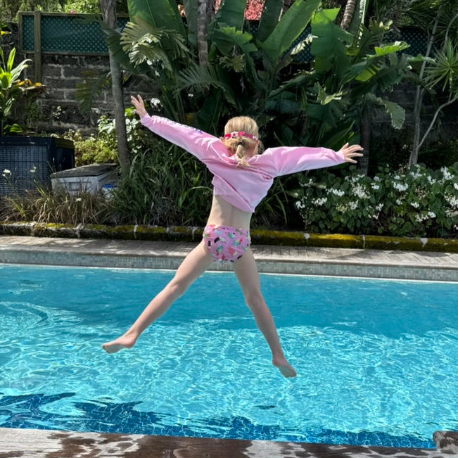 Child in pink shirt and shorts jumping into a pool with lush greenery in the background