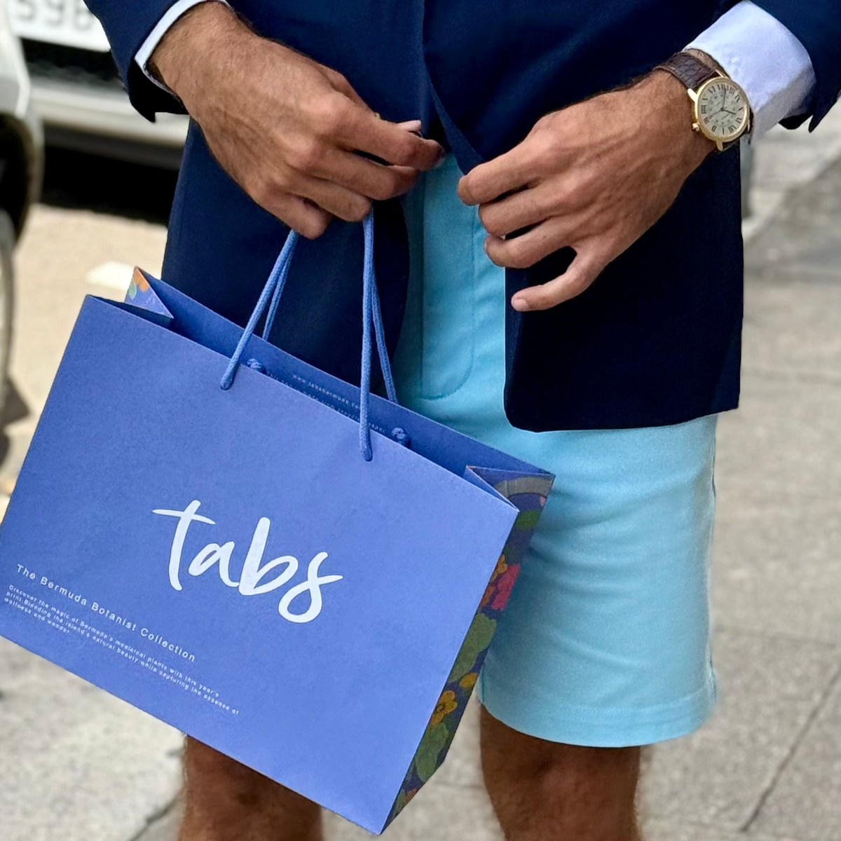 Person wearing a navy blazer and light blue shorts, holding a blue 'tabs' shopping bag.