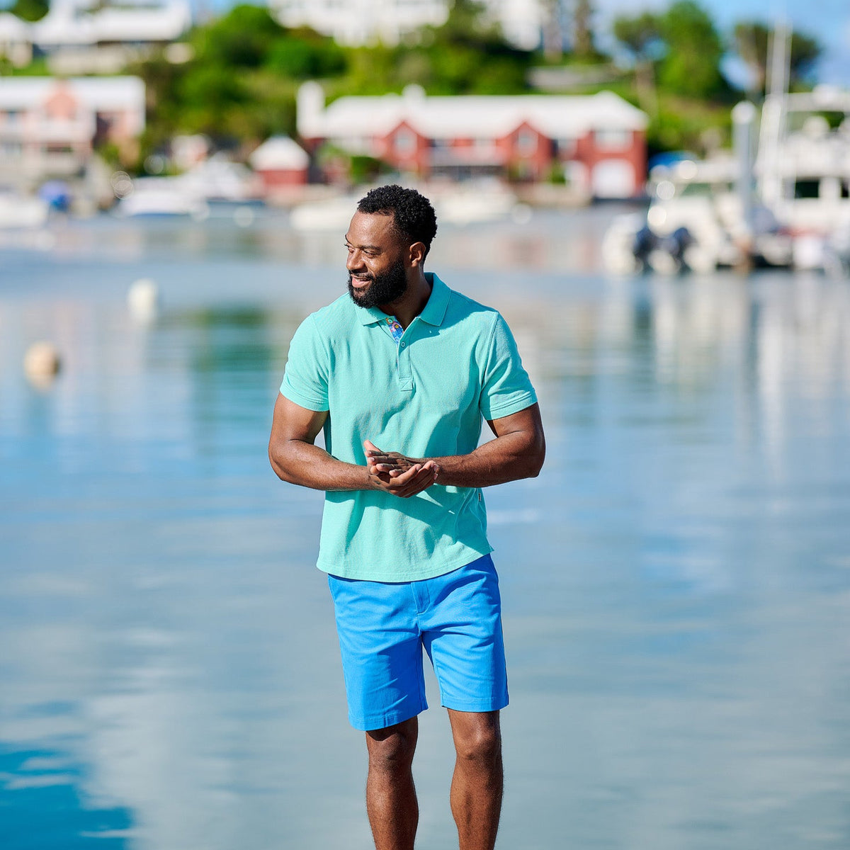 Man in turquoise shirt and blue shorts standing by a waterfront with boats and buildings in the background.