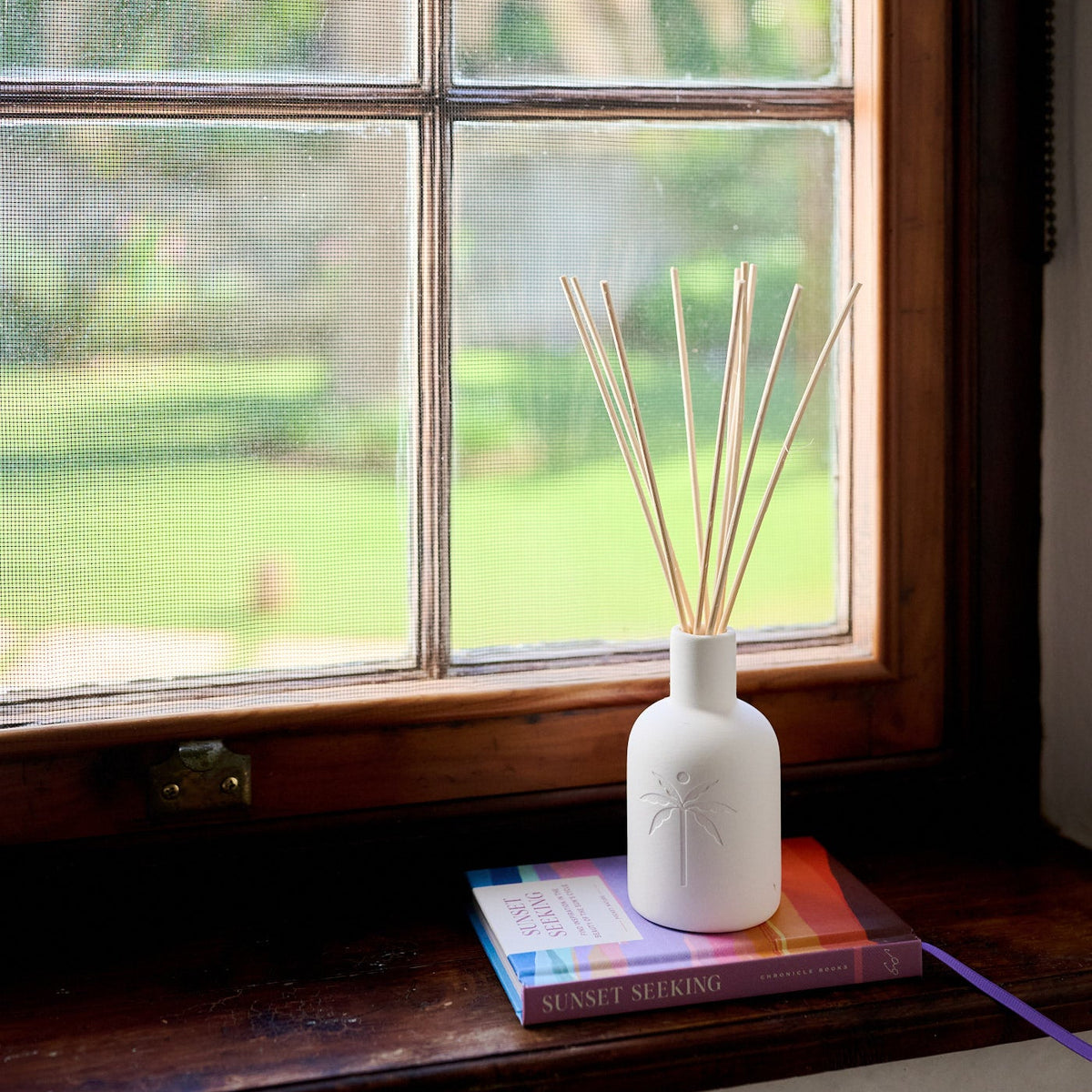 White diffuser with reeds on a windowsill with a view of greenery outside.
