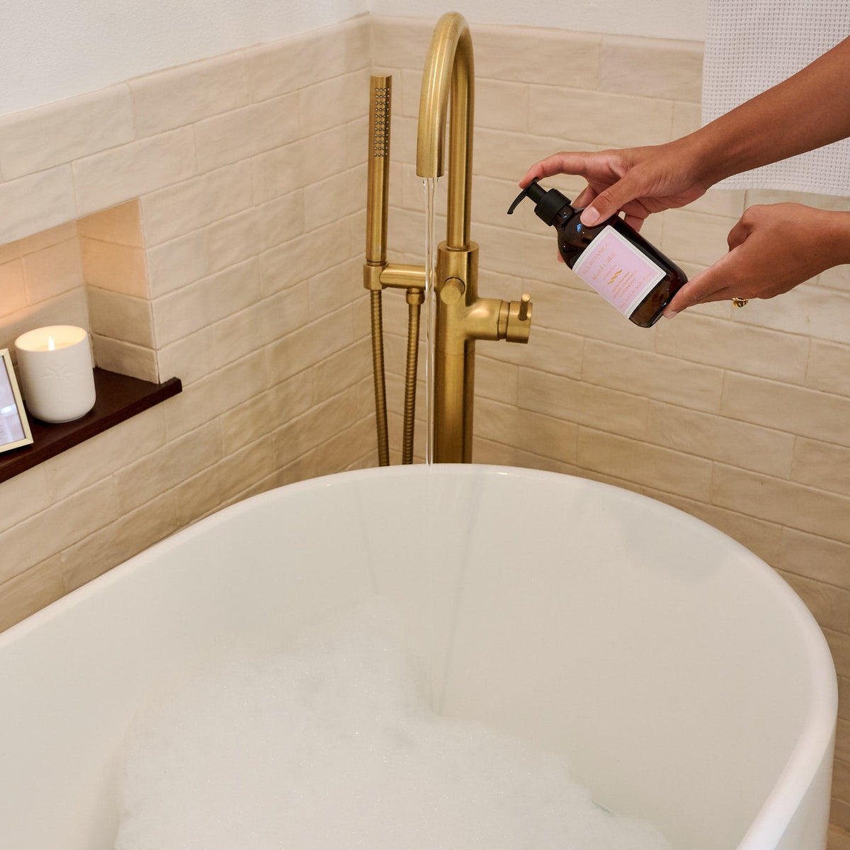 Person pouring a liquid from a bottle into a bathtub with a gold faucet and decorative items.