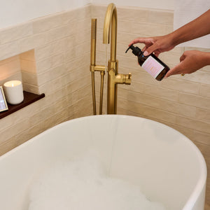 Person pouring a liquid from a bottle into a bathtub with a gold faucet and decorative items.