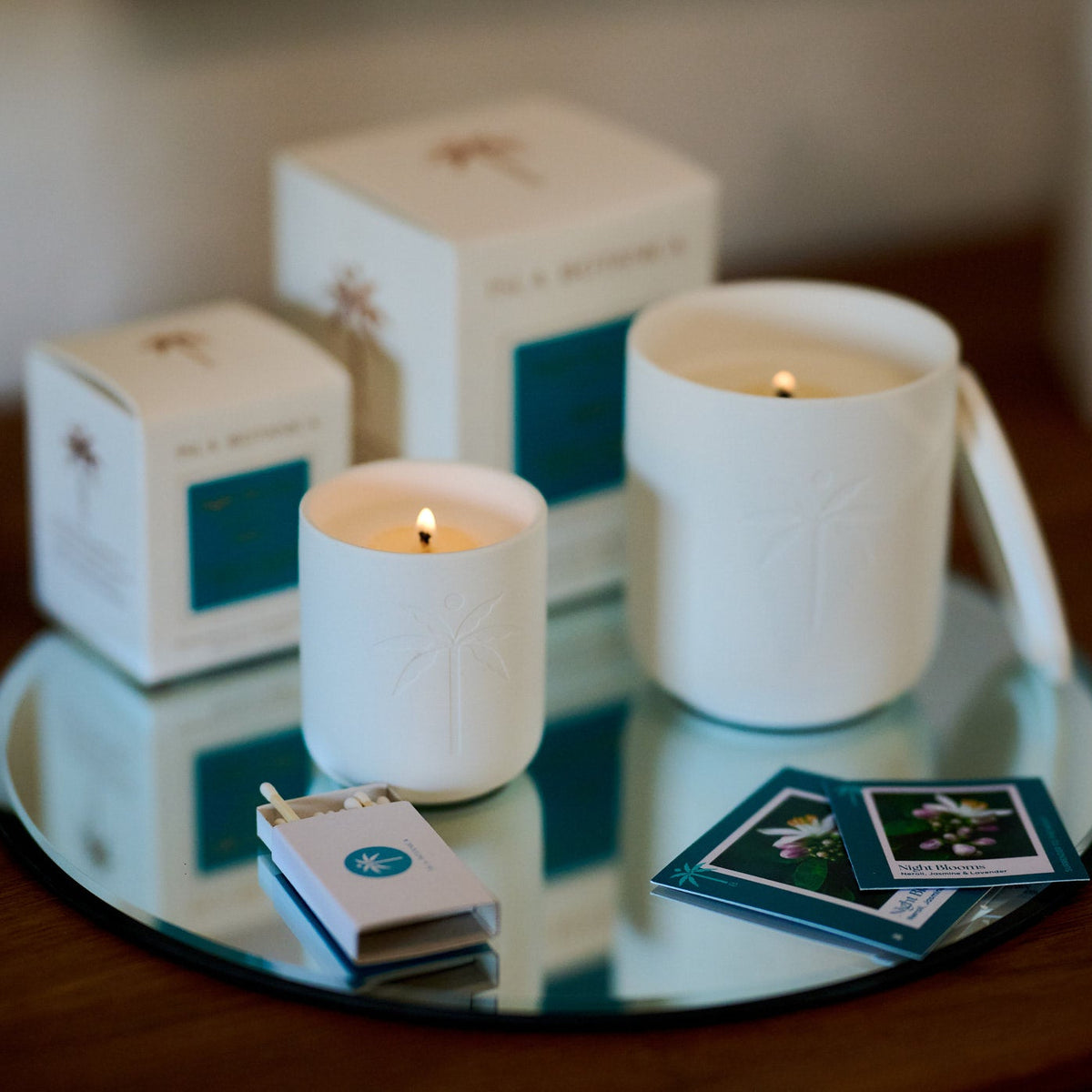 Two white candles on a reflective surface with boxes and cards in the background