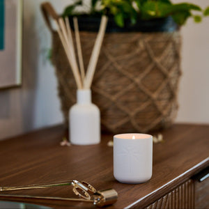 White candle and diffuser on a wooden surface with a plant in the background