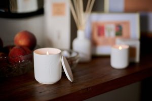 Two white candles on a wooden surface with peaches and decorative items in the background.
