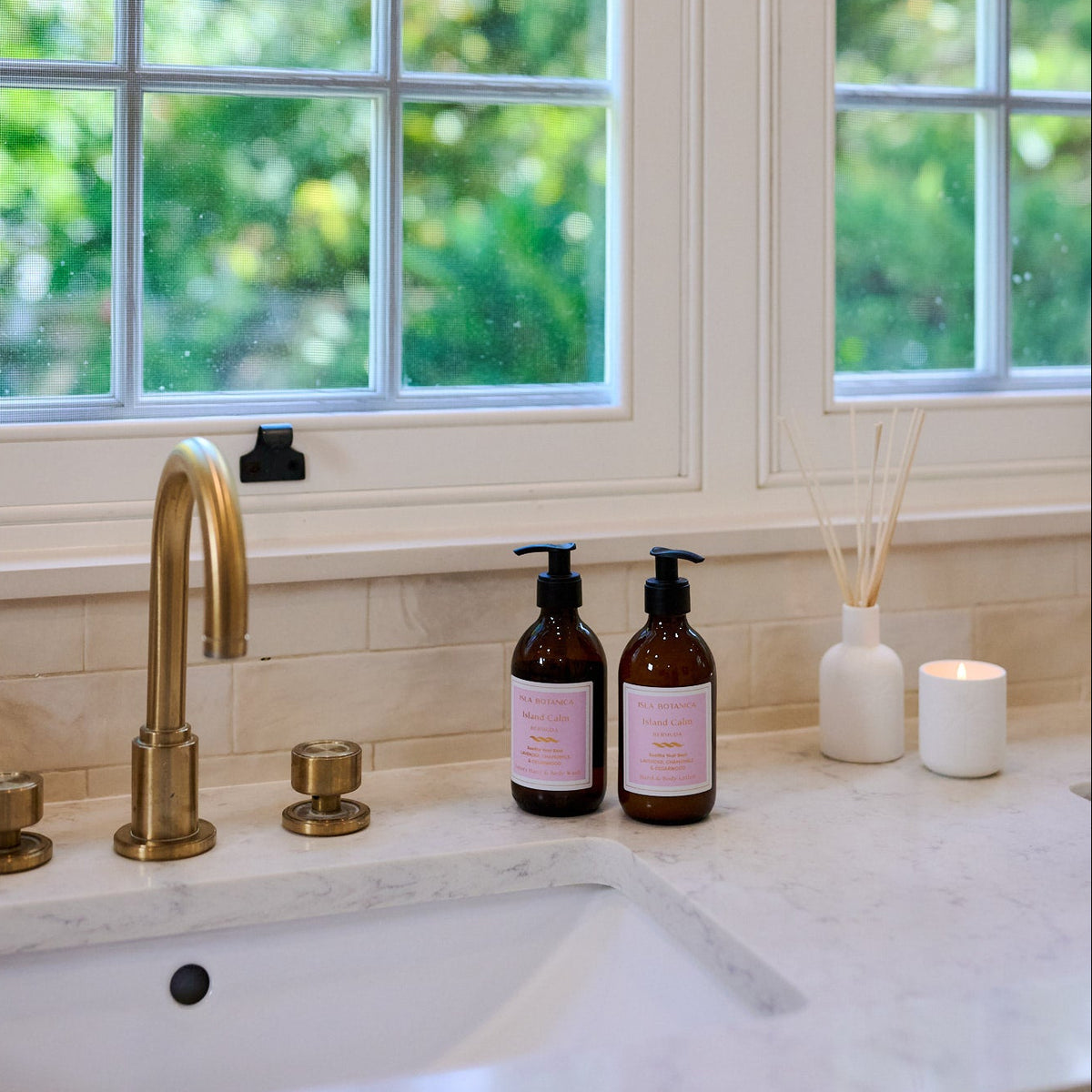 Kitchen sink area with gold faucet, bottles, and a candle on a windowsill.