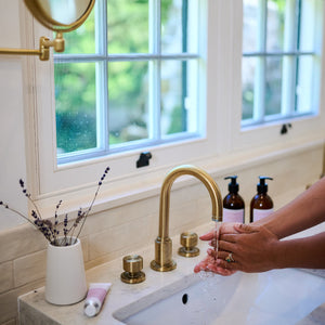 Person washing hands under a gold faucet in a bathroom with a window in the background.
