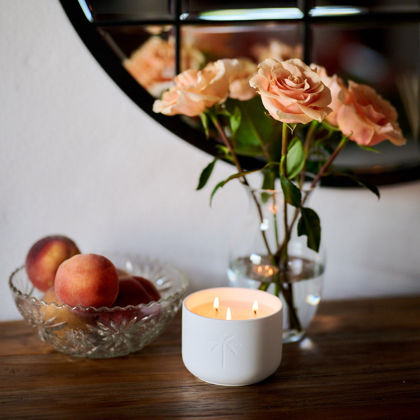 Vase of pink roses, bowl of peaches, and white candle on a wooden surface with a mirror in the background.