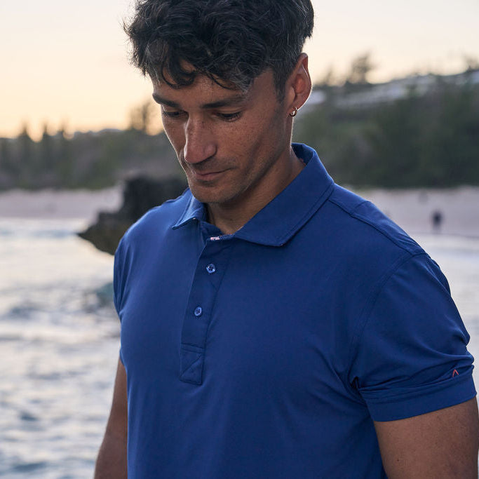 Man wearing a blue polo shirt standing by a body of water with a sunset or sunrise in the background.