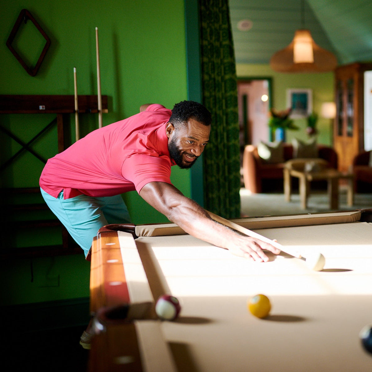 Man in a pink shirt playing pool in a room with green walls and furniture.
