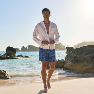 Man standing on a beach wearing a white shirt and blue shorts with a scenic background.