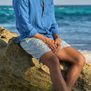 Person sitting on a rock by the ocean wearing a blue shirt and patterned shorts.