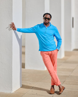 Man wearing a blue shirt and pink pants leaning against a white wall.