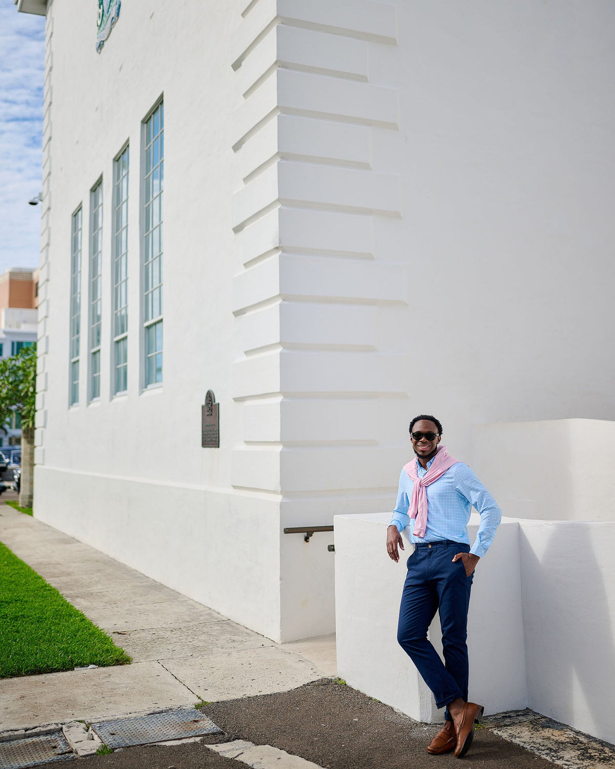 Man leaning against a white building wall wearing sunglasses and a light blue shirt.