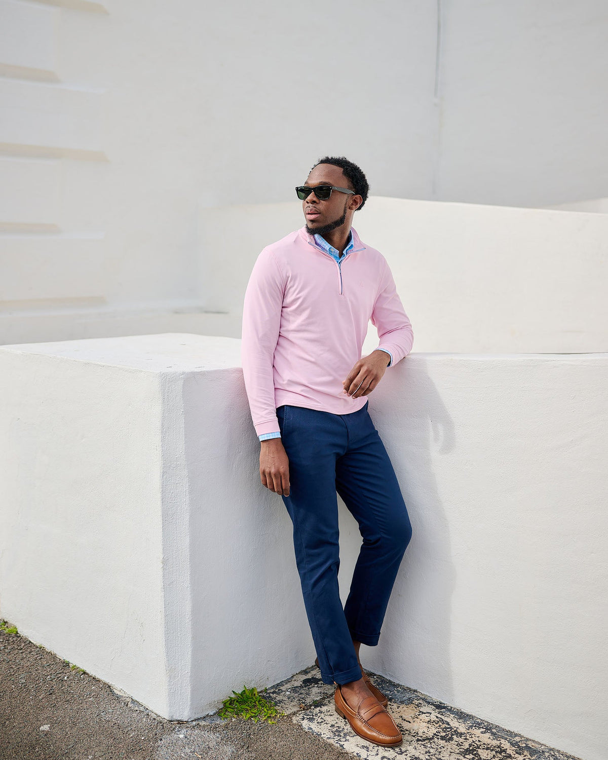 Man in pink sweater and blue pants leaning against a white wall.