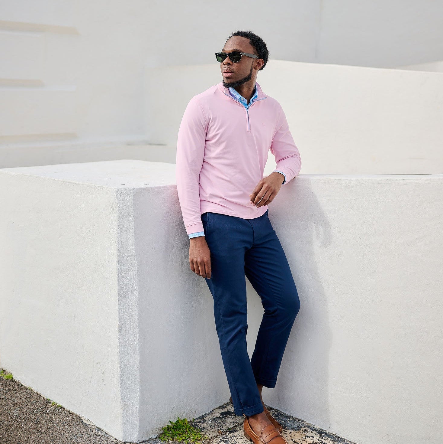 Man in pink sweater and blue pants leaning against a white wall.
