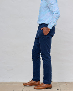 Man wearing a light blue patterned shirt, dark blue pants, and brown shoes against a white wall.